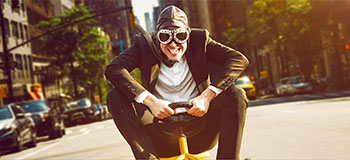 Person in a suit riding a small toy vehicle on a city street, with tall buildings and traffic in the background under bright sunlight.