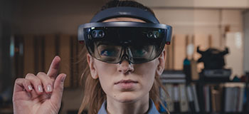 Person wearing an augmented reality headset and raising one hand, with shelves and office equipment visible in the background.