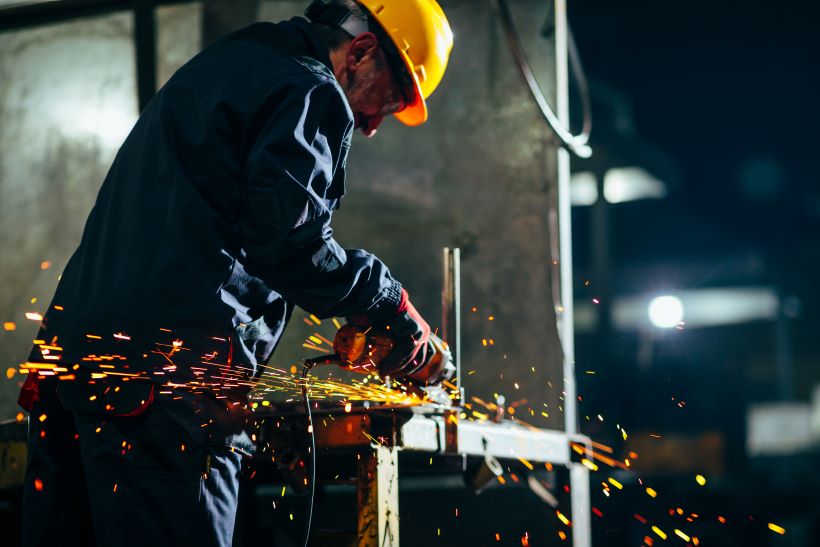Worker wearing a safety helmet using a power tool to grind metal, producing bright sparks in an industrial setting.