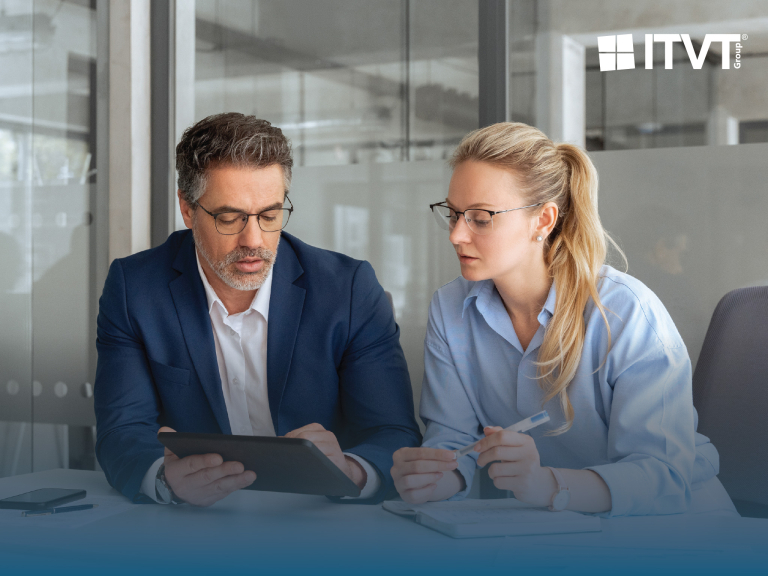 Two business people, a man in a blue suit and a woman in a light blue shirt, both wearing glasses, are checking information on a tablet in an office environment. The ITVT Group logo is visible in the upper right corner.