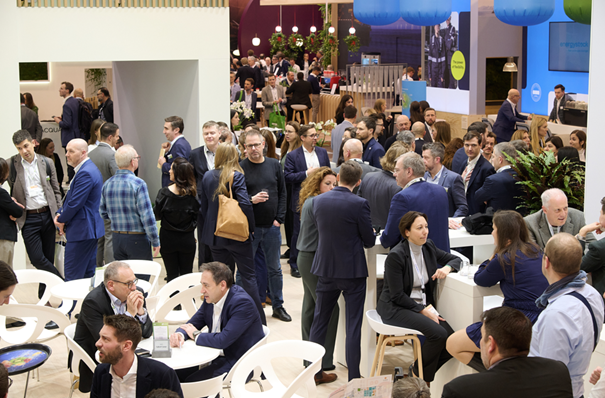 A busy ITVT event scene at a trade‑fair venue, showing a large group of attendees networking, conversing, and gathering around tables and booths. The space is well‑lit with modern exhibition stands, seating areas, plants, and digital displays visible in the background. The atmosphere is active and professional, with many people engaged in discussions throughout the booth area.