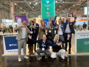 A group of people posing energetically for a group photo at an ITVT exhibition booth. The booth features bright signage, including banners for ITVT, d.velop, and 365 Stadtwerk. A large green display in the background highlights ‘Autonomous Agents + Copilot.’ The scene is set in a busy trade‑fair hall with lights, stands, and visitors visible throughout the venue.
